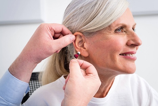 Eine ältere Frau bekommt ein modernes Hörgerät am Ohr angepasst und lächelt zufrieden.